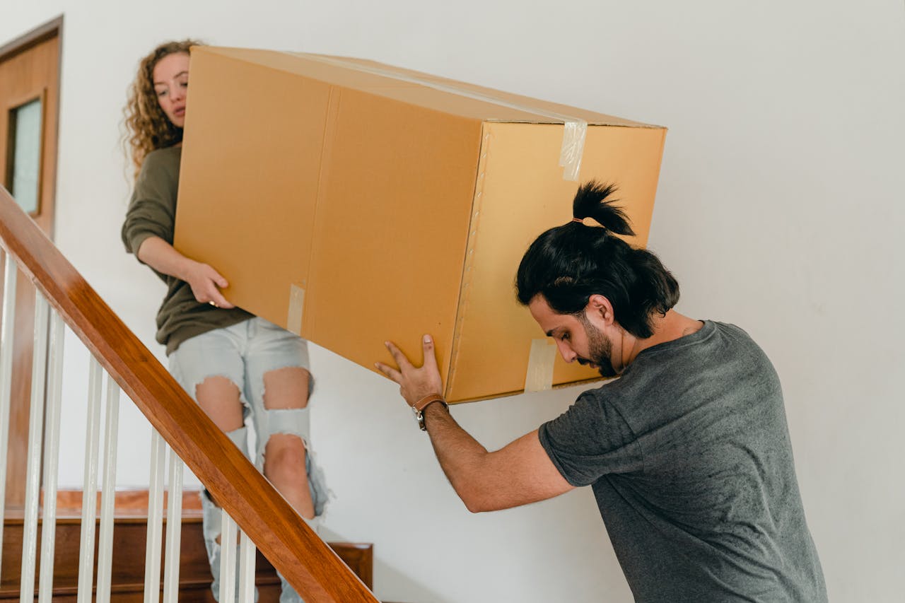 A young couple carries a large cardboard box up a wooden staircase, symbolizing the moving process.